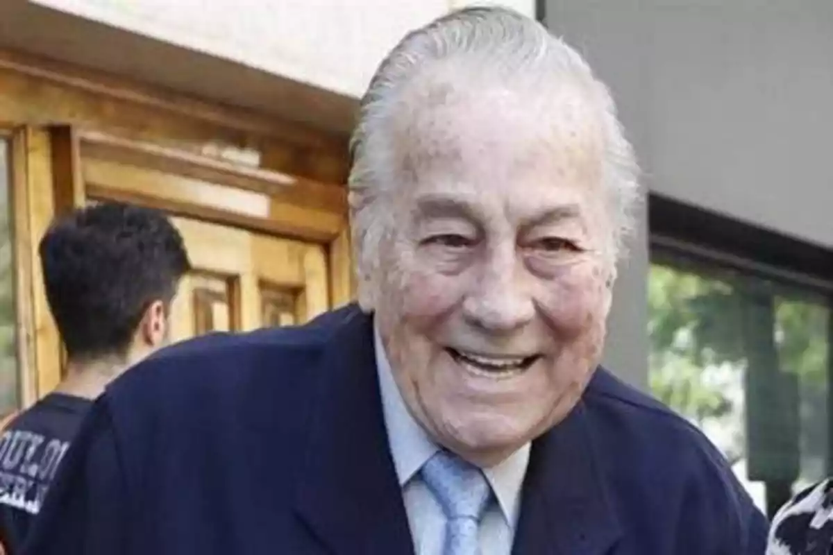 Un hombre mayor sonriendo, vestido con traje y corbata, con un fondo de una puerta de madera y una ventana.