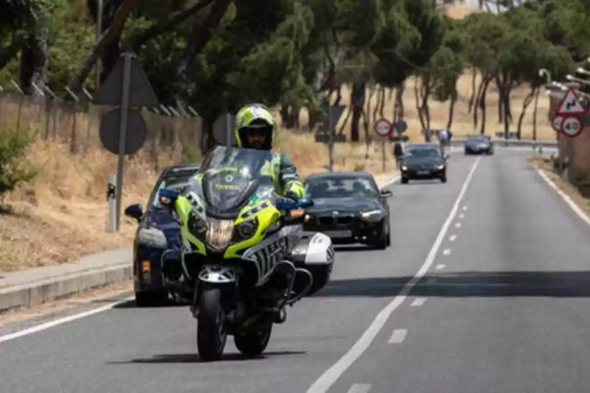 Un oficial de policía en una motocicleta patrulla una carretera, seguido por varios coches, en un entorno rural con árboles y señales de tráfico.