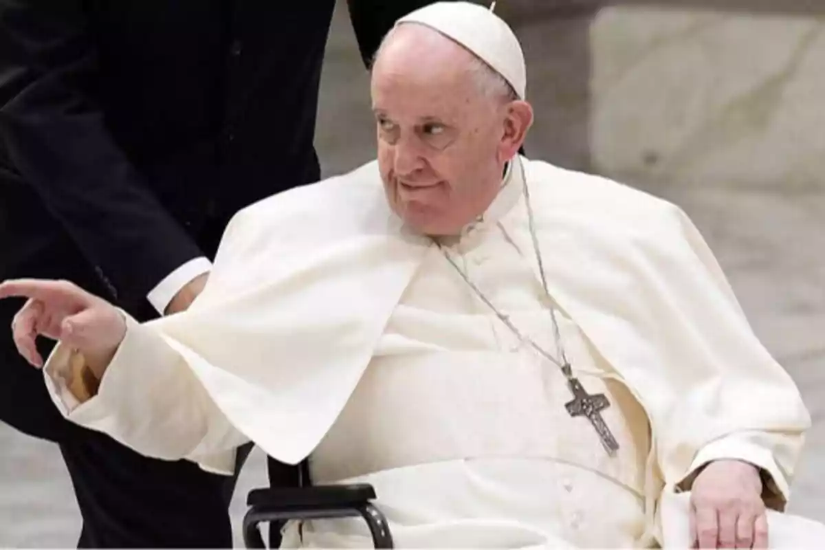 Persona vestida con atuendo religioso blanco y sombrero, sentada en una silla de ruedas y señalando con la mano.