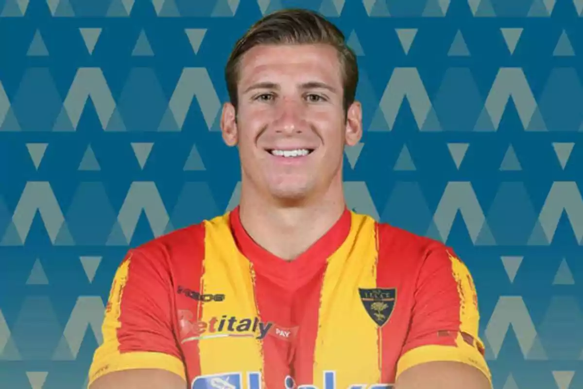 Un jugador de fútbol con una camiseta roja y amarilla sonríe frente a un fondo azul con patrones geométricos.