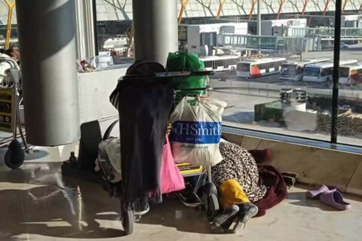 Una persona durmiendo en el suelo junto a un carrito de equipaje lleno de bolsas y ropa en un aeropuerto.