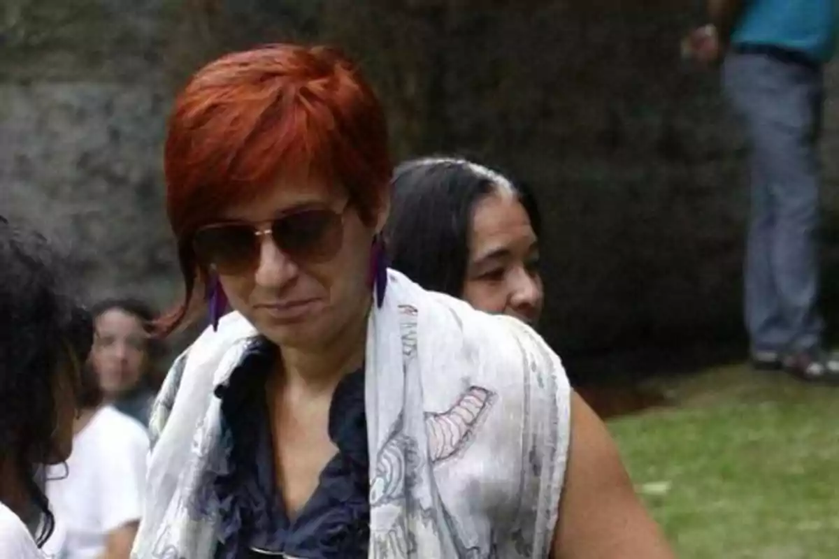 Una mujer con cabello corto y rojo, usando gafas de sol y un chal blanco con dibujos, está al aire libre con otras personas en el fondo.