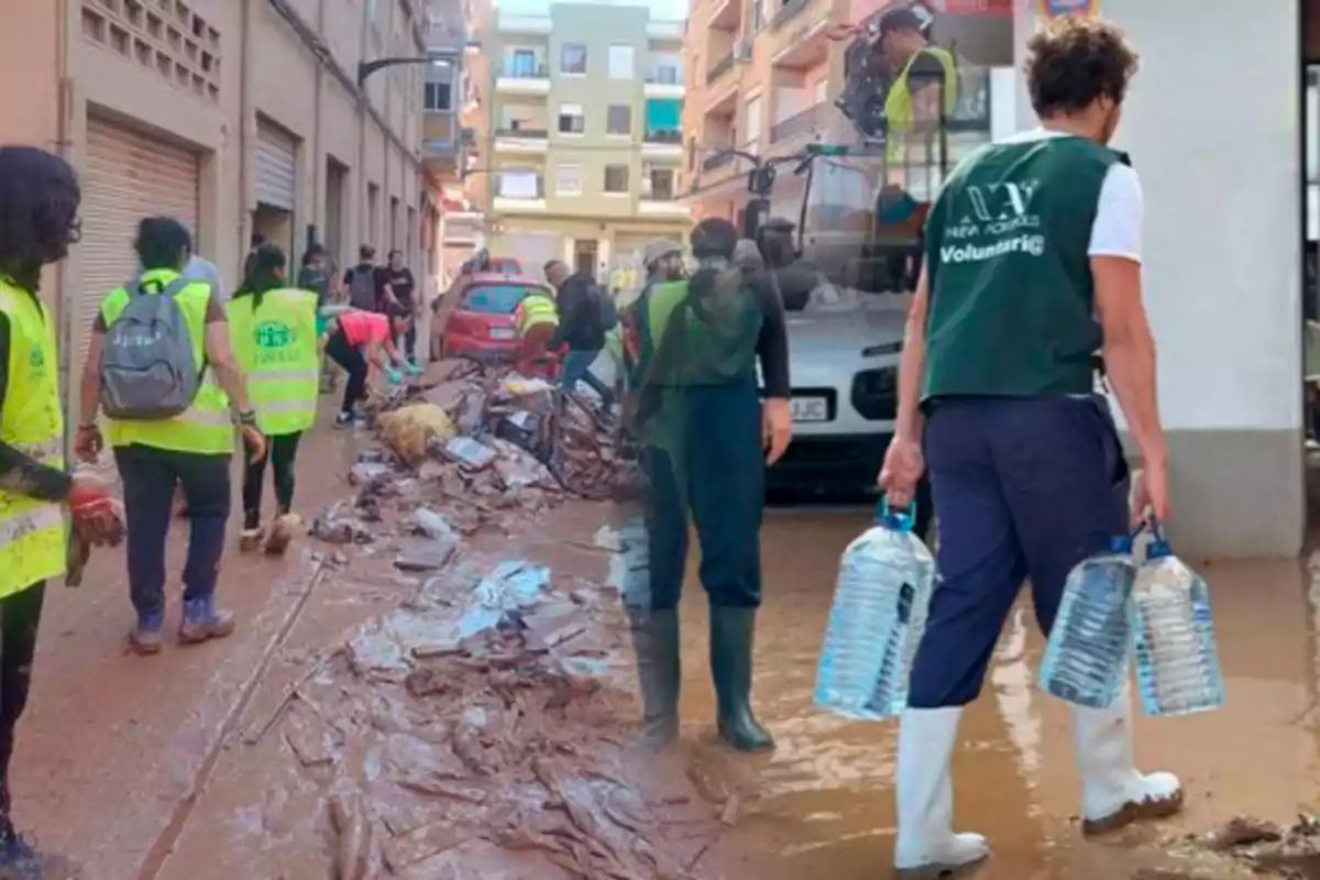 Voluntarios con chalecos y botas de goma ayudan a limpiar una calle llena de escombros y barro tras una inundación, mientras una persona lleva garrafas de agua.