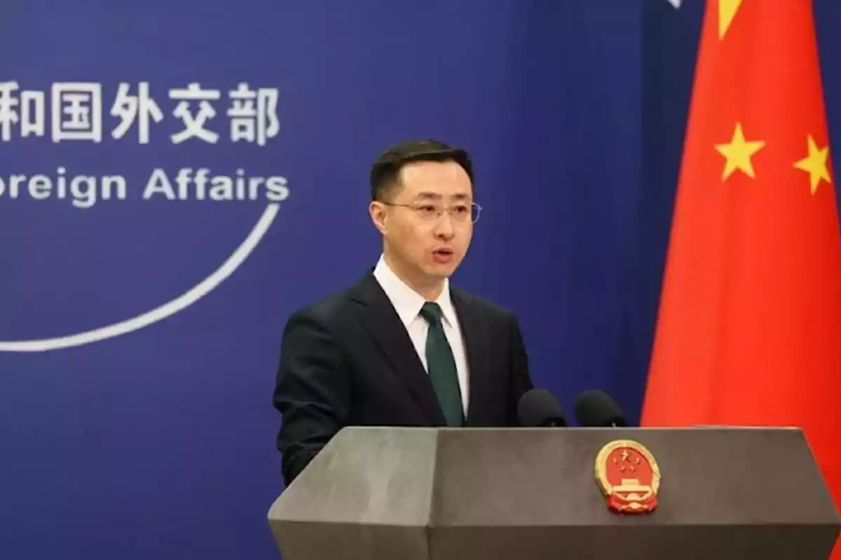 Un hombre en un podio con el emblema de China, hablando frente a un fondo azul con texto en chino e inglés y una bandera china a la derecha.