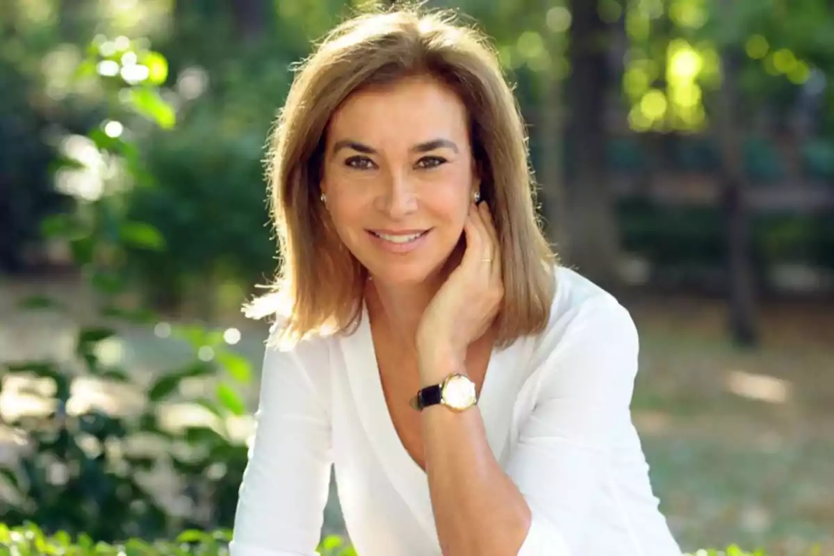 Una mujer sonriente con cabello castaño claro y una blusa blanca, posando en un entorno al aire libre con vegetación de fondo.