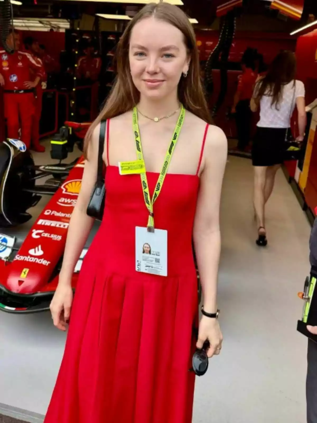 Alejandra de Hannover con un vestido rojo y una acreditación colgada en el cuello está de pie frente a un coche de carreras en un garaje. Alejandra de Hannover con un vestido rojo y una acreditación colgada en el cuello está de pie frente a un coche de carreras en un garaje.