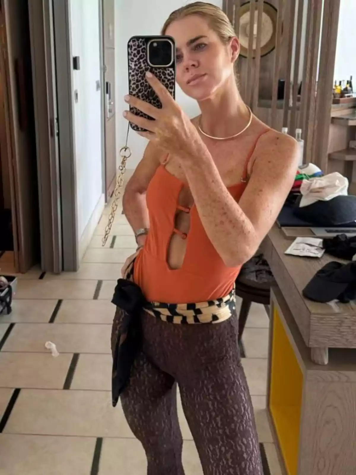 Amelia Bono se toma una selfie frente a un espejo usando un traje de baño naranja y leggings oscuros con un cinturón estampado, en un ambiente interior moderno. Amelia Bono se toma una selfie frente a un espejo usando un traje de baño naranja y leggings oscuros con un cinturón estampado, en un ambiente interior moderno.