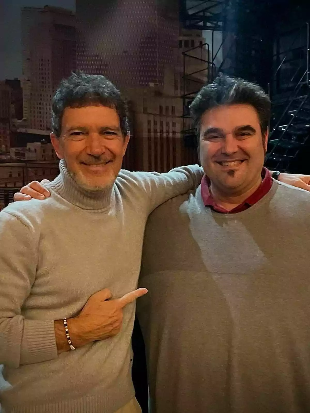 Antonio Banderas y Txema Cariñena sonríen mientras uno de ellos señala al otro con el brazo alrededor de su hombro, ambos visten suéteres y están en un entorno urbano.