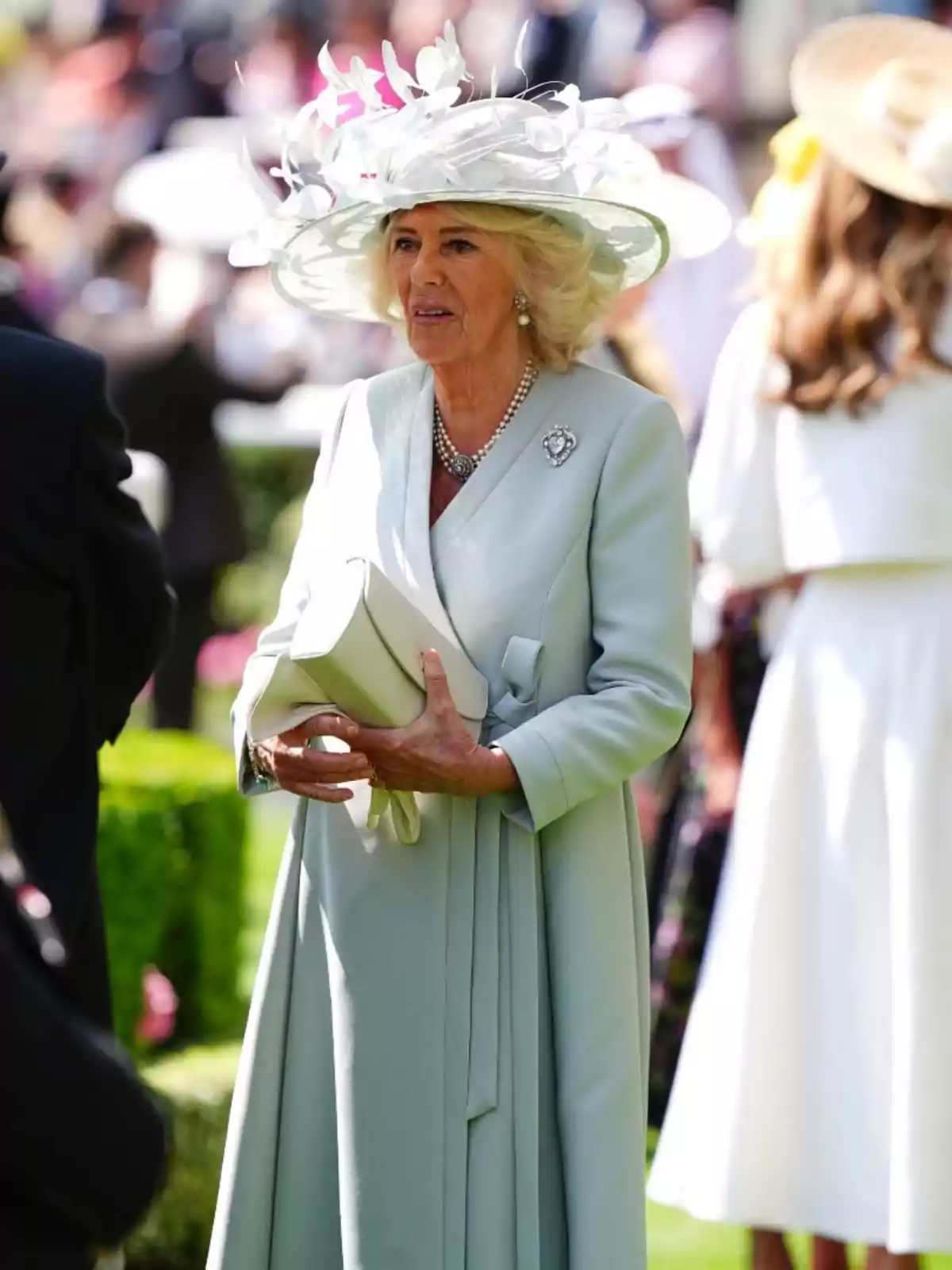 La reina Camila elegantemente vestida con abrigo celeste y sombrero decorado con flores blancas sosteniendo un bolso en un evento al aire libre La reina Camila elegantemente vestida con abrigo celeste y sombrero decorado con flores blancas sosteniendo un bolso en un evento al aire libre