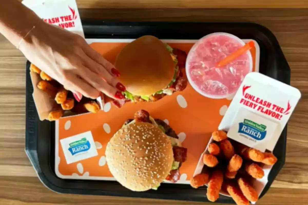 Una bandeja con hamburguesas, papas fritas, una bebida rosada y un aderezo, con una mano tomando una de las hamburguesas. Una bandeja con hamburguesas, papas fritas, una bebida rosada y un aderezo, con una mano tomando una de las hamburguesas.