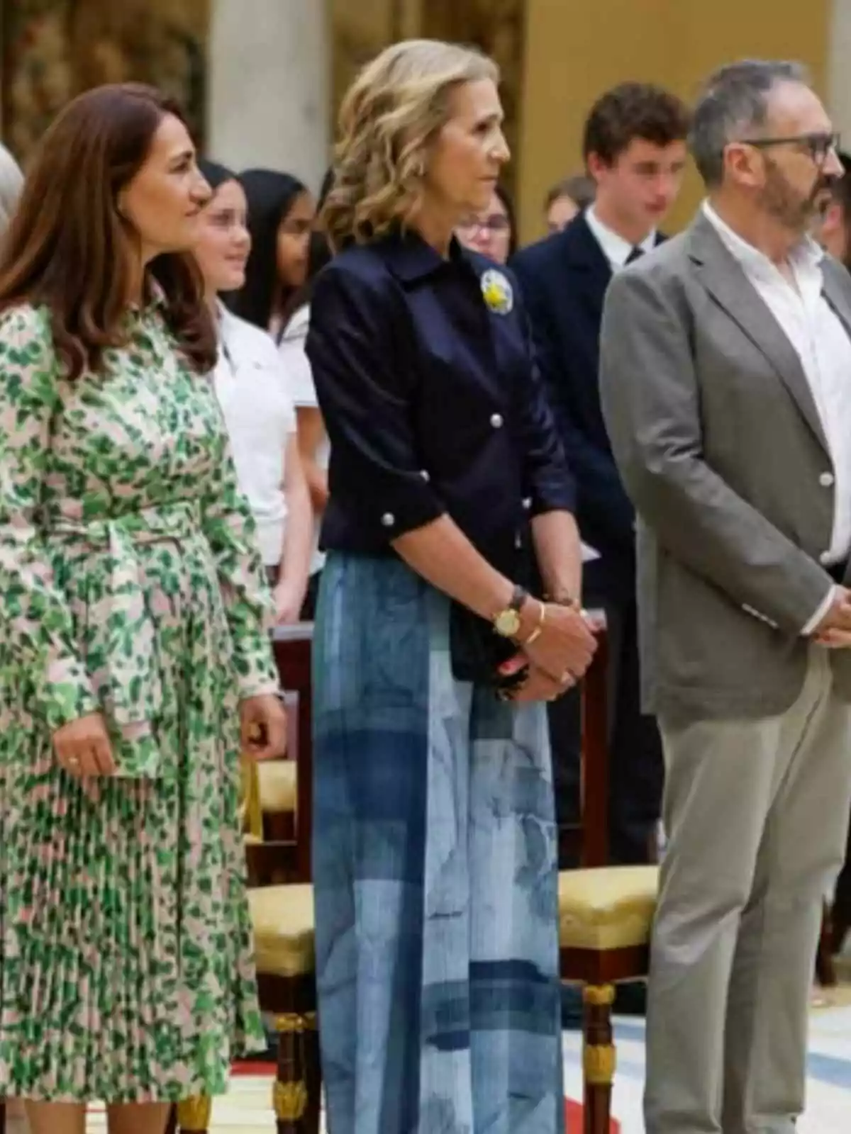 Infanta Elena and two other people standing at a formal event, two women and a man, with several people in the background. Infanta Elena and two other people standing at a formal event, two women and a man, with several people in the background.