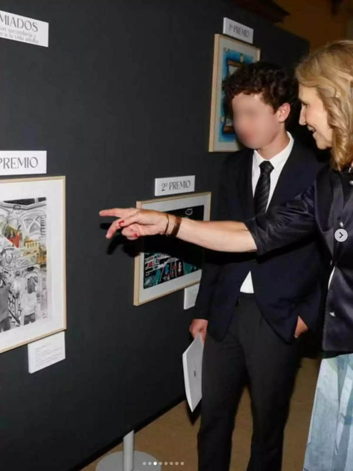 Infanta Elena points to a work of art at an exhibition while a young man in a suit watches her. Infanta Elena points to a work of art at an exhibition while a young man in a suit watches her.