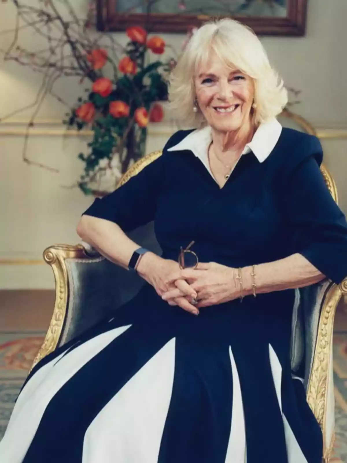 La reina Camila con vestido azul oscuro con detalles blancos está sentada en una silla elegante, sonriendo frente a un fondo decorado con flores naranjas. La reina Camila con vestido azul oscuro con detalles blancos está sentada en una silla elegante, sonriendo frente a un fondo decorado con flores naranjas.