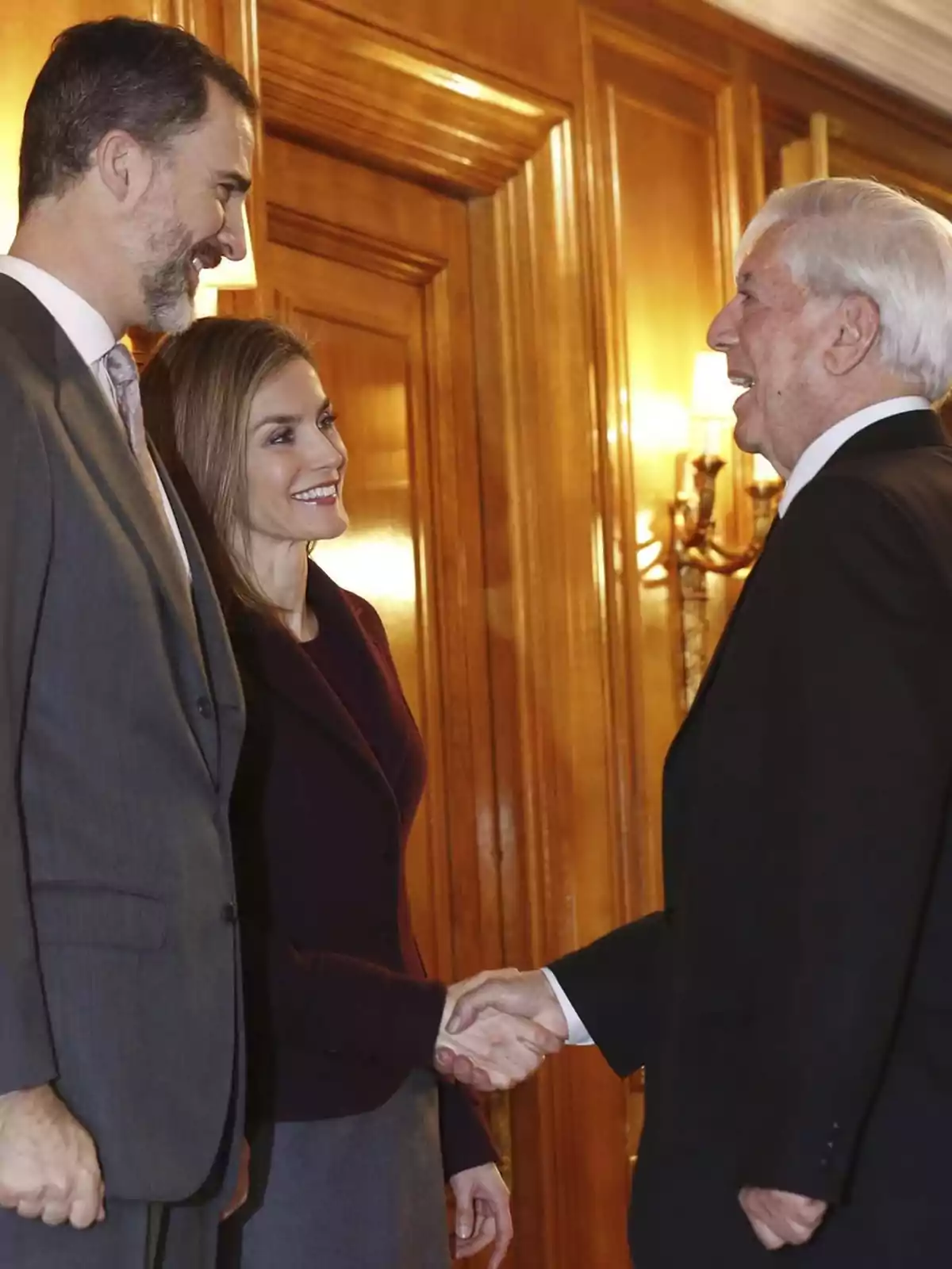 Felipe VI, la reina Letizia y Mario Vargas Llosa se saludan cordialmente en un entorno formal con paredes de madera y luces cálidas. Felipe VI, la reina Letizia y Mario Vargas Llosa se saludan cordialmente en un entorno formal con paredes de madera y luces cálidas.