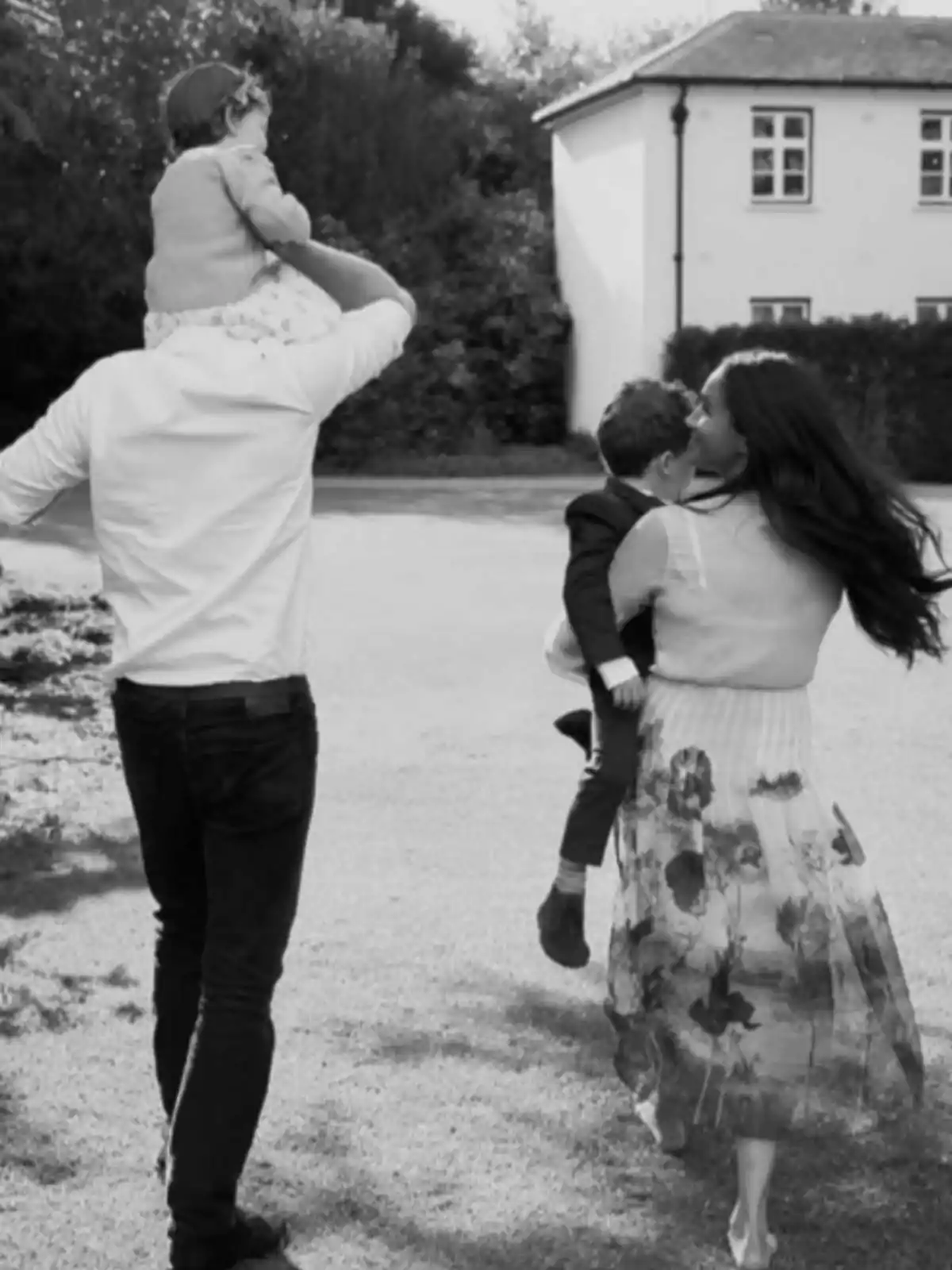 Una familia caminando al aire libre, con un hombre llevando a una niña sobre sus hombros y una mujer cargando a un niño en brazos, mientras se dirigen hacia una casa. Una familia caminando al aire libre, con un hombre llevando a una niña sobre sus hombros y una mujer cargando a un niño en brazos, mientras se dirigen hacia una casa.
