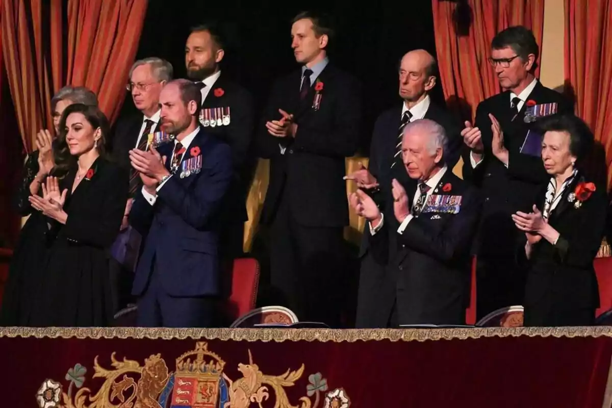 La familia real británica vestidas formalmente aplaude en un evento, con cortinas rojas de fondo y un escudo decorativo en primer plano.