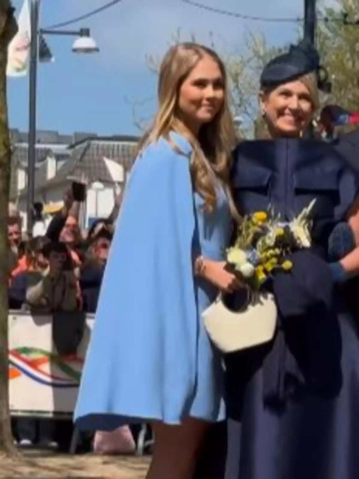 Dos mujeres posan juntas al aire libre, una con un vestido azul claro y la otra con un atuendo azul oscuro y un sombrero, mientras personas en el fondo toman fotos. Dos mujeres posan juntas al aire libre, una con un vestido azul claro y la otra con un atuendo azul oscuro y un sombrero, mientras personas en el fondo toman fotos.