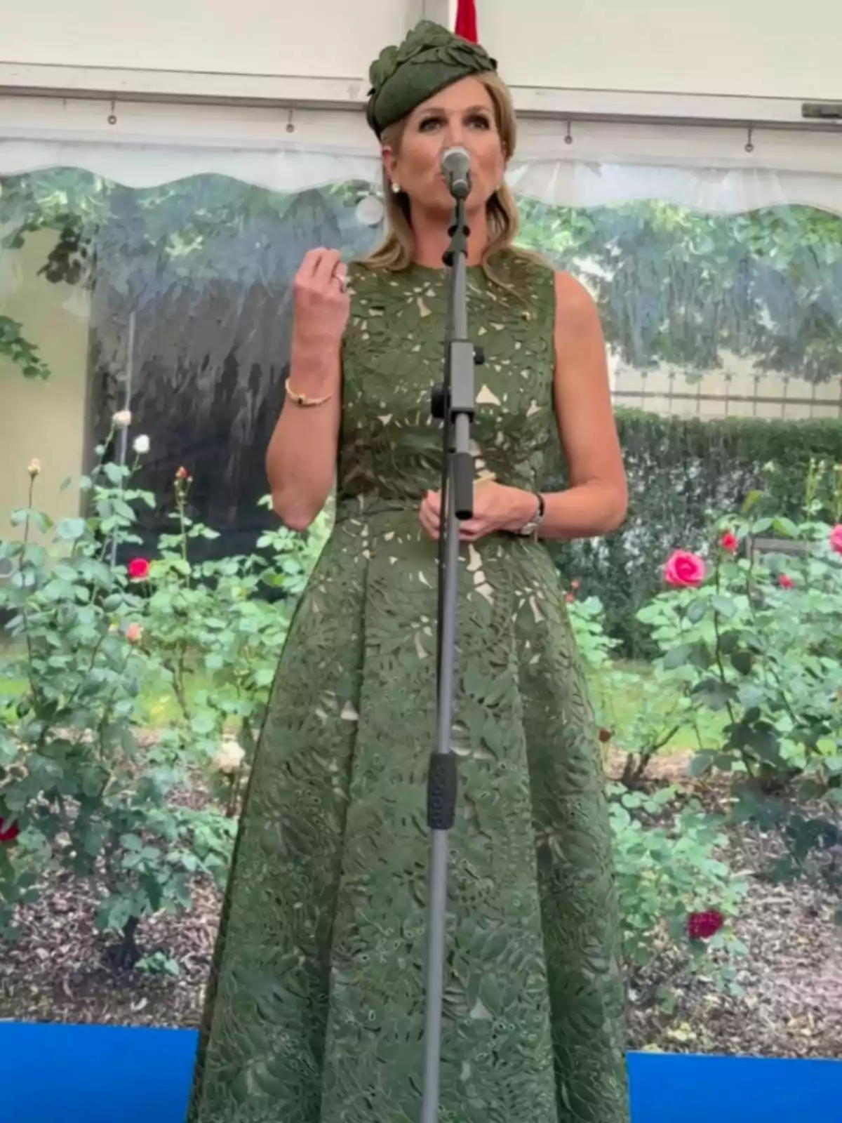Máxima de Holanda con vestido y sombrero verde hablando frente a un micrófono en un jardín con rosas al fondo Máxima de Holanda con vestido y sombrero verde hablando frente a un micrófono en un jardín con rosas al fondo