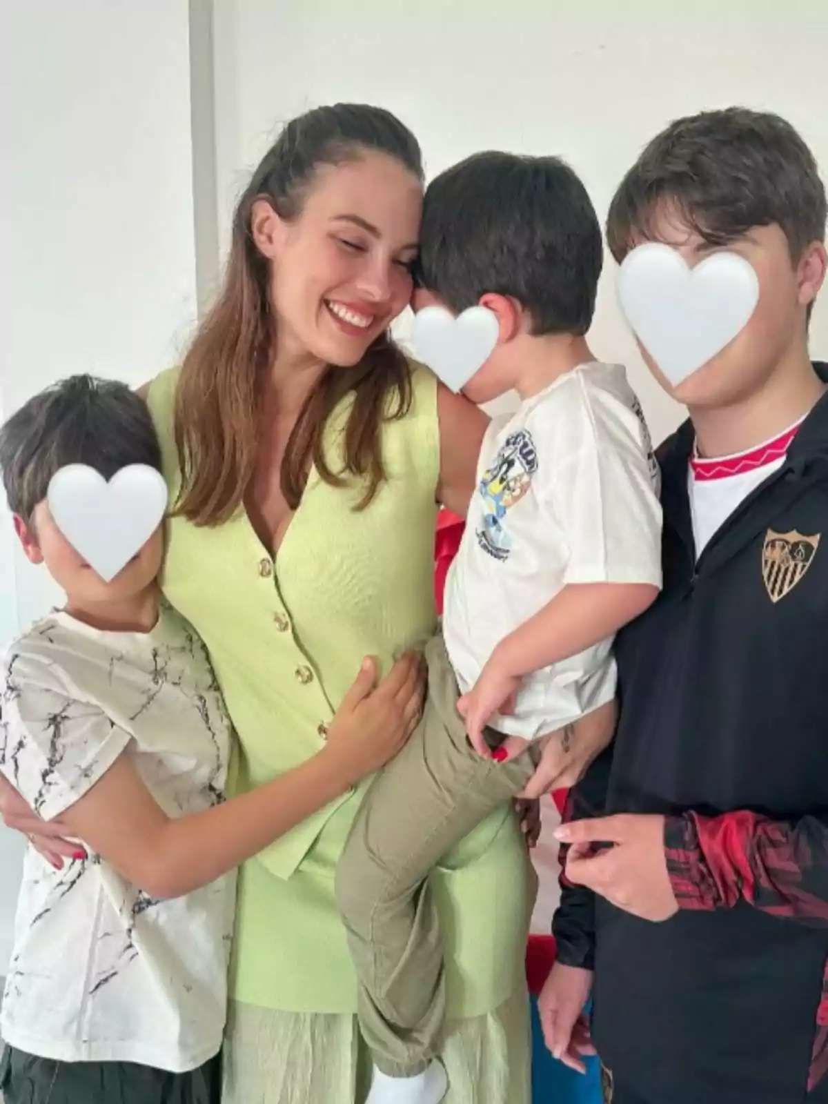 Jessica Bueno sonriente sostiene a un niño pequeño mientras está rodeada de otros dos niños, todos con caras cubiertas por corazones blancos. Jessica Bueno sonriente sostiene a un niño pequeño mientras está rodeada de otros dos niños, todos con caras cubiertas por corazones blancos.