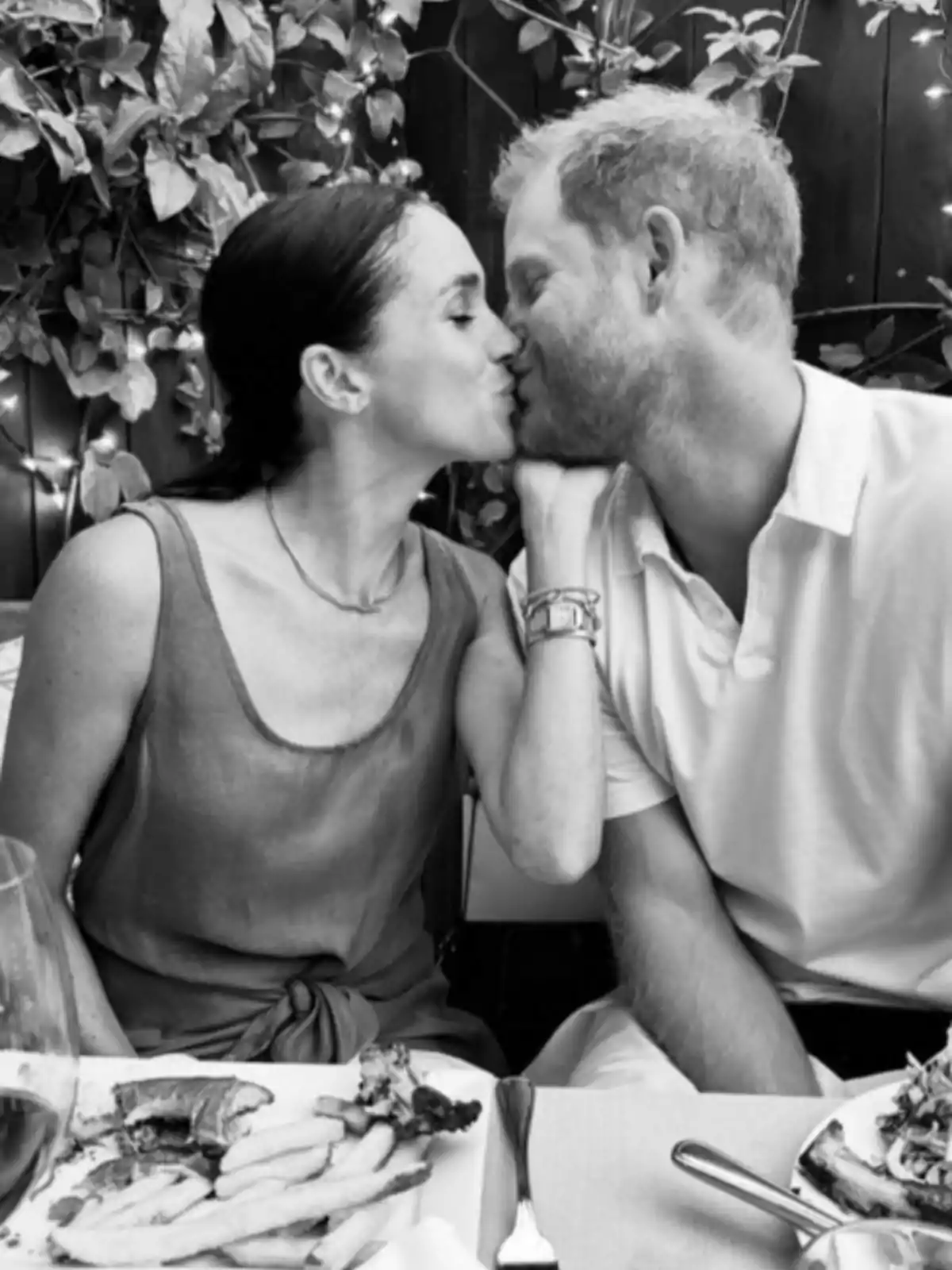 Meghan Markle y el príncipe Harry compartiendo un beso en una mesa con comida y luces decorativas en el fondo. Meghan Markle y el príncipe Harry compartiendo un beso en una mesa con comida y luces decorativas en el fondo.