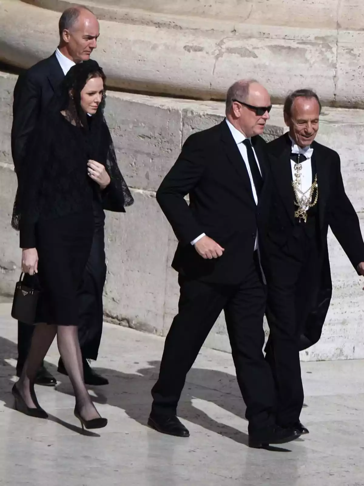A group of people dressed formally walks next to a stone building. A group of people dressed formally walks next to a stone building.