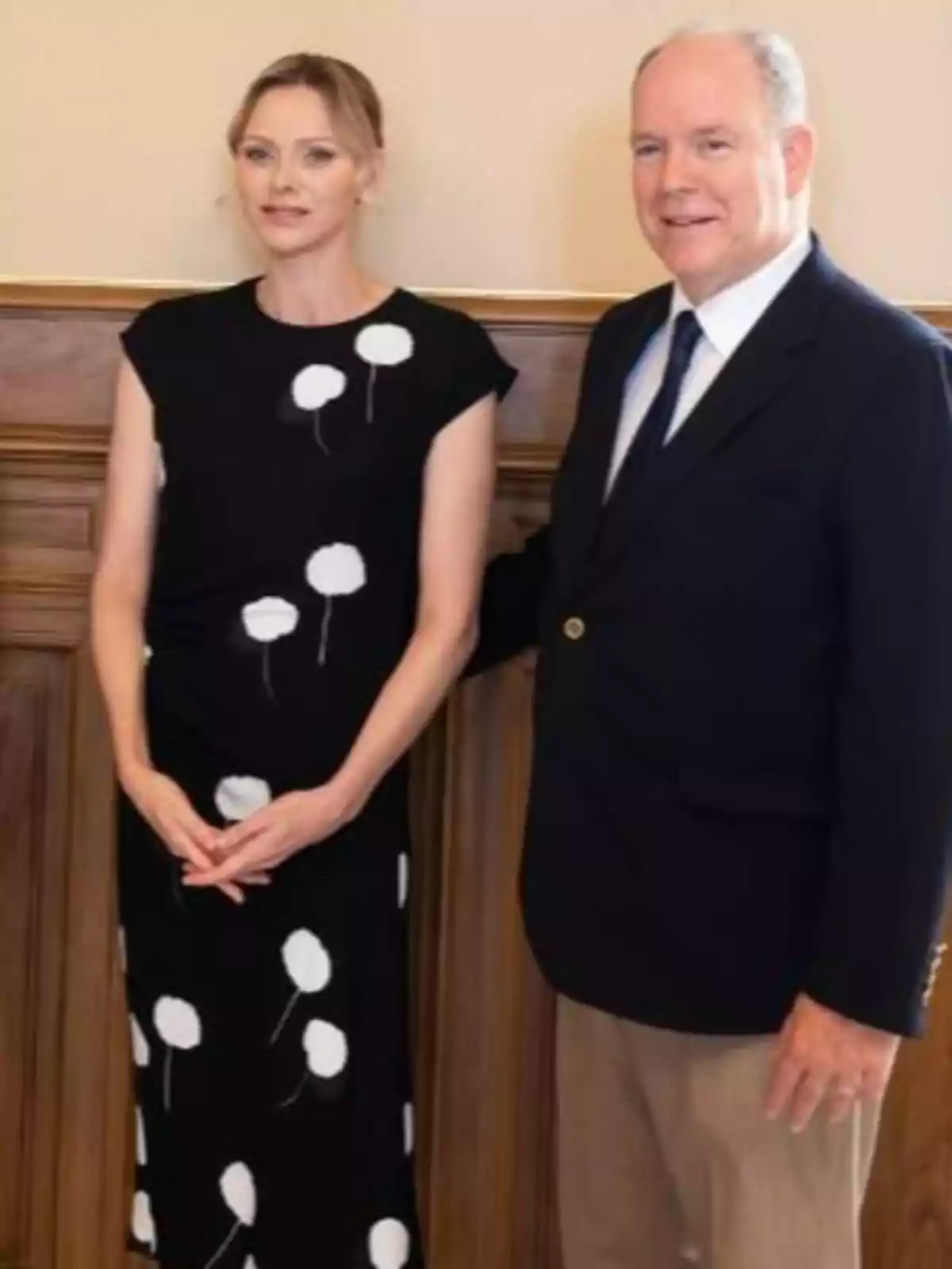 Charlene in a black dress with white polka dots and Albert of Monaco in a dark jacket and light pants pose together in an elegant interior.