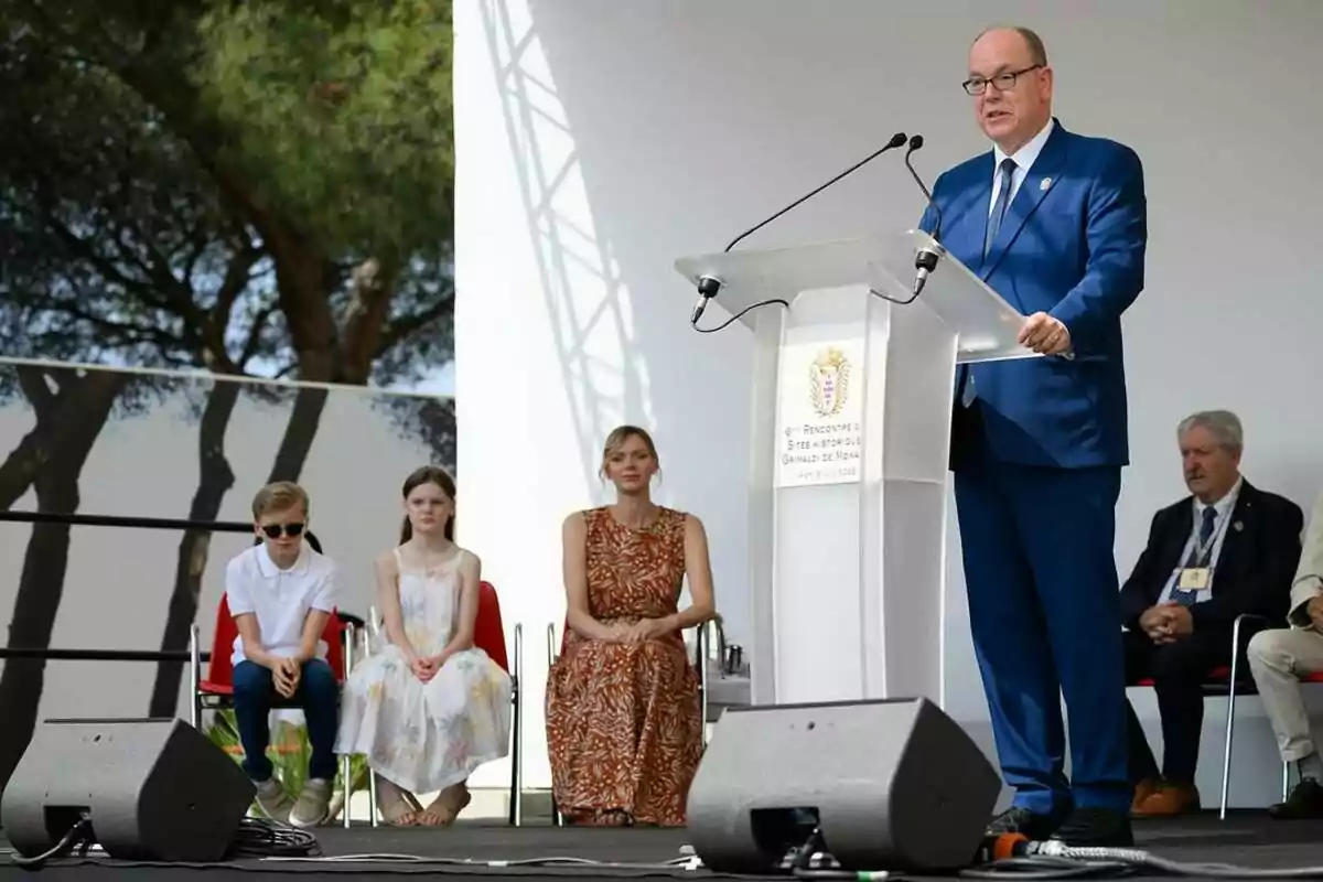 Alberto de Mónaco con traje azul habla en un podio durante un evento al aire libre mientras varias personas, incluyendo dos niños y una mujer, están sentados detrás de él Alberto de Mónaco con traje azul habla en un podio durante un evento al aire libre mientras varias personas, incluyendo dos niños y una mujer, están sentados detrás de él