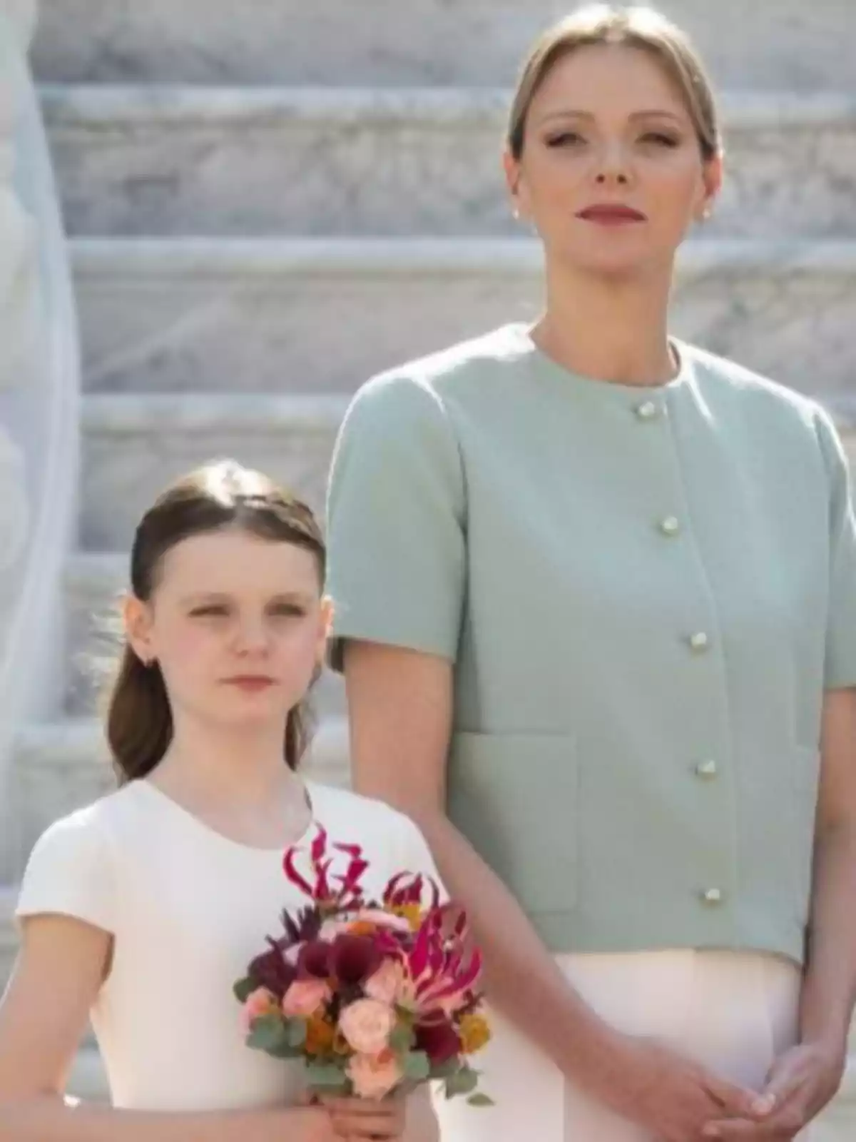 Charlene y Gabriella de Mónaco posan juntas en una escalera de mármol, la niña sostiene un ramo de flores y ambas miran al frente con expresión seria Charlene y Gabriella de Mónaco posan juntas en una escalera de mármol, la niña sostiene un ramo de flores y ambas miran al frente con expresión seria