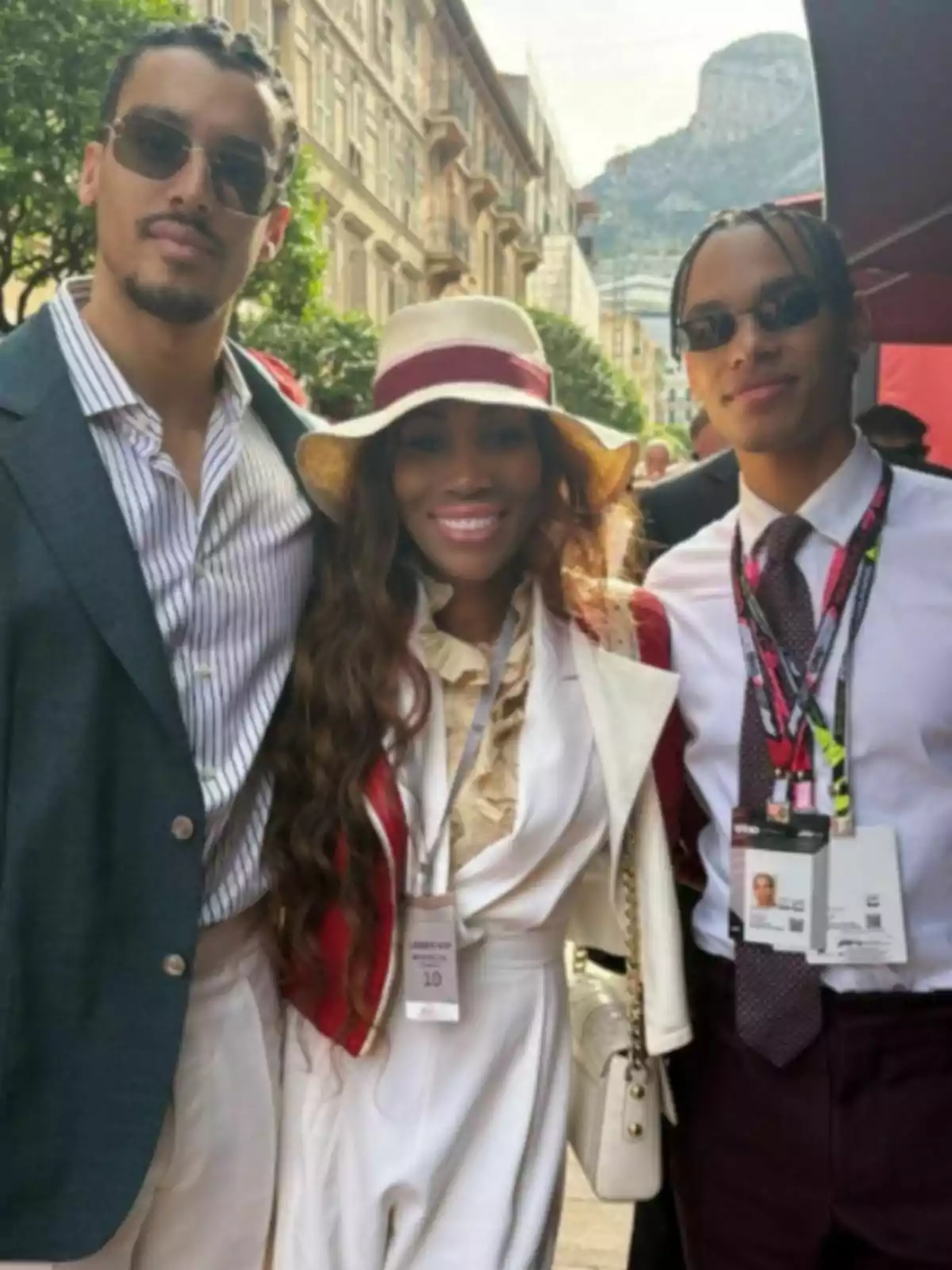 Three people pose smiling outdoors on an elegant street; a woman in the center wears a wide-brimmed hat, and the two men beside her are wearing sunglasses and formal clothing. In the background, there are buildings and a mountain. Three people pose smiling outdoors on an elegant street; a woman in the center wears a wide-brimmed hat, and the two men beside her are wearing sunglasses and formal clothing. In the background, there are buildings and a mountain.