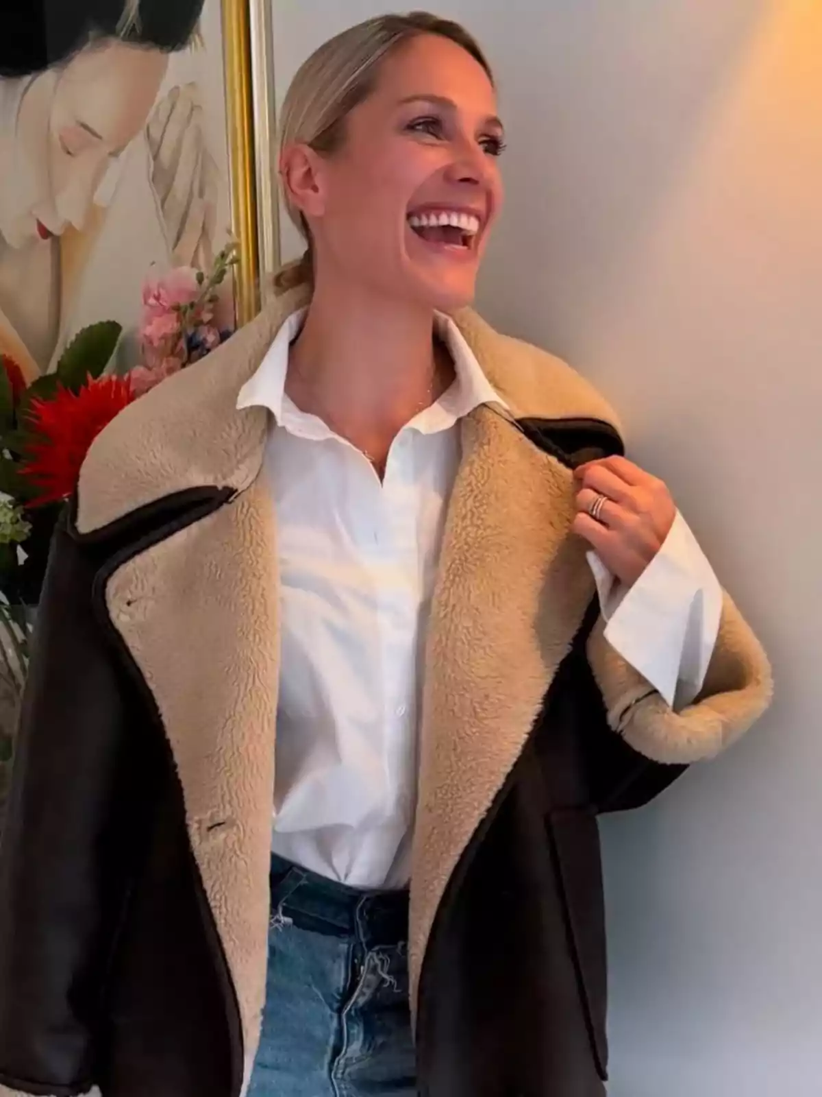 Pascalle Paerel sonriente con una chaqueta de piel sintética y camisa blanca, de pie junto a una pintura y flores. Pascalle Paerel sonriente con una chaqueta de piel sintética y camisa blanca, de pie junto a una pintura y flores.