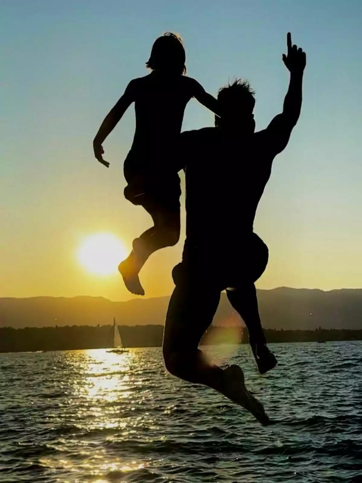 Una silueta de Perico Durán y un niño saltando juntos al agua al atardecer con el sol y un velero en el horizonte.