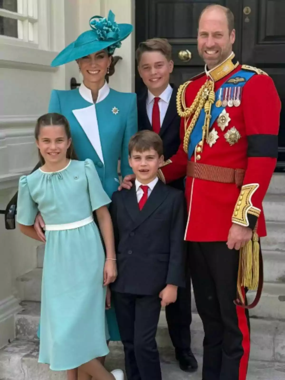 An elegantly dressed family poses smiling at the entrance of a building, with the man wearing a red uniform with medals and the woman a blue dress with a matching hat, accompanied by three well-groomed children. An elegantly dressed family poses smiling at the entrance of a building, with the man wearing a red uniform with medals and the woman a blue dress with a matching hat, accompanied by three well-groomed children.