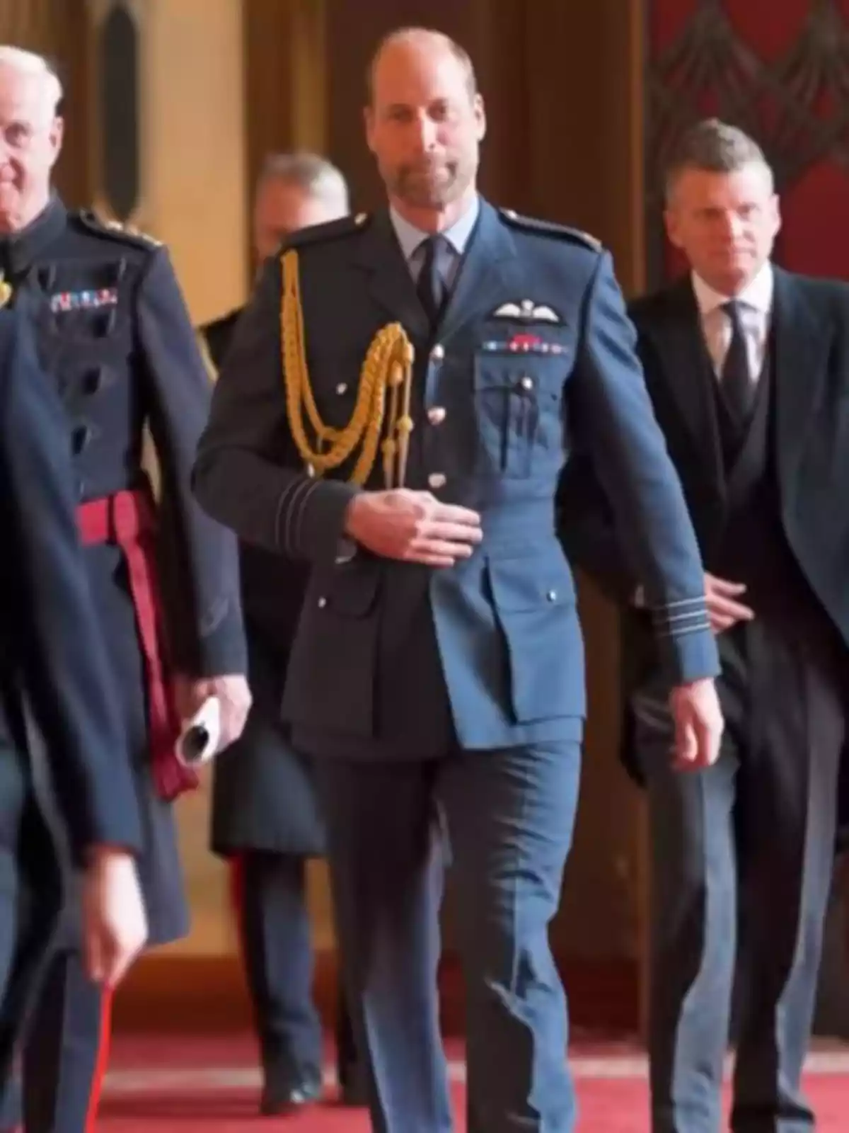 El príncipe Guillermo con uniforme militar azul camina por un pasillo alfombrado acompañado de otros hombres vestidos de manera formal El príncipe Guillermo con uniforme militar azul camina por un pasillo alfombrado acompañado de otros hombres vestidos de manera formal