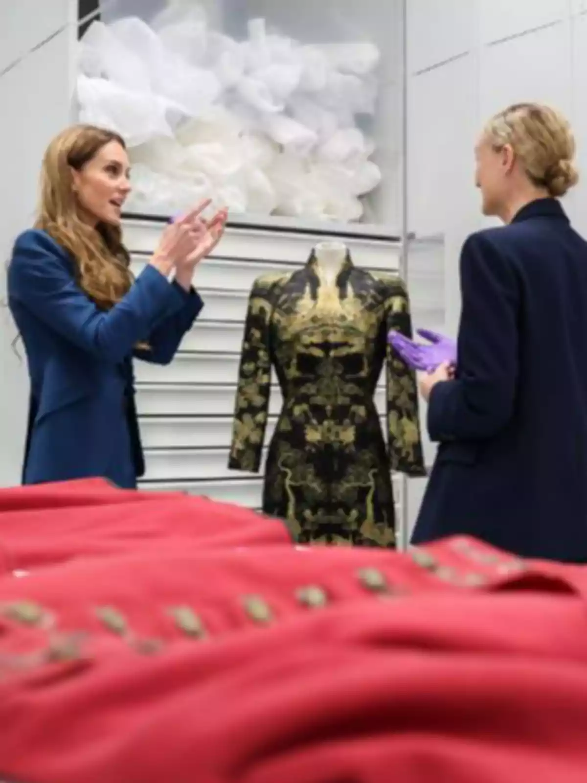 Kate Middleton y otra mujer conversan frente a un maniquí con un vestido negro y dorado en una sala donde hay varias prendas rojas sobre una mesa Kate Middleton y otra mujer conversan frente a un maniquí con un vestido negro y dorado en una sala donde hay varias prendas rojas sobre una mesa