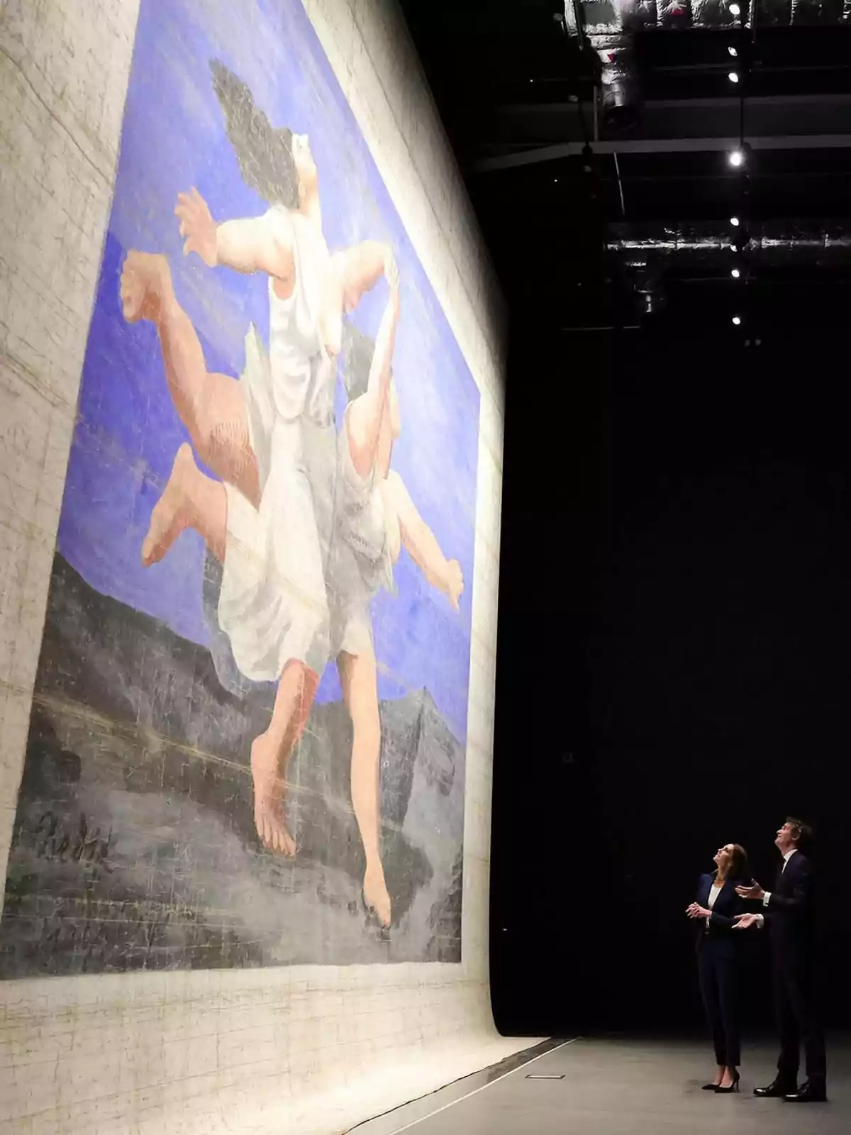 Kate Middleton y un hombre observan un gran telón pintado con la imagen de dos figuras femeninas en movimiento sobre un fondo azul y montañoso dentro de un espacio oscuro y amplio Kate Middleton y un hombre observan un gran telón pintado con la imagen de dos figuras femeninas en movimiento sobre un fondo azul y montañoso dentro de un espacio oscuro y amplio