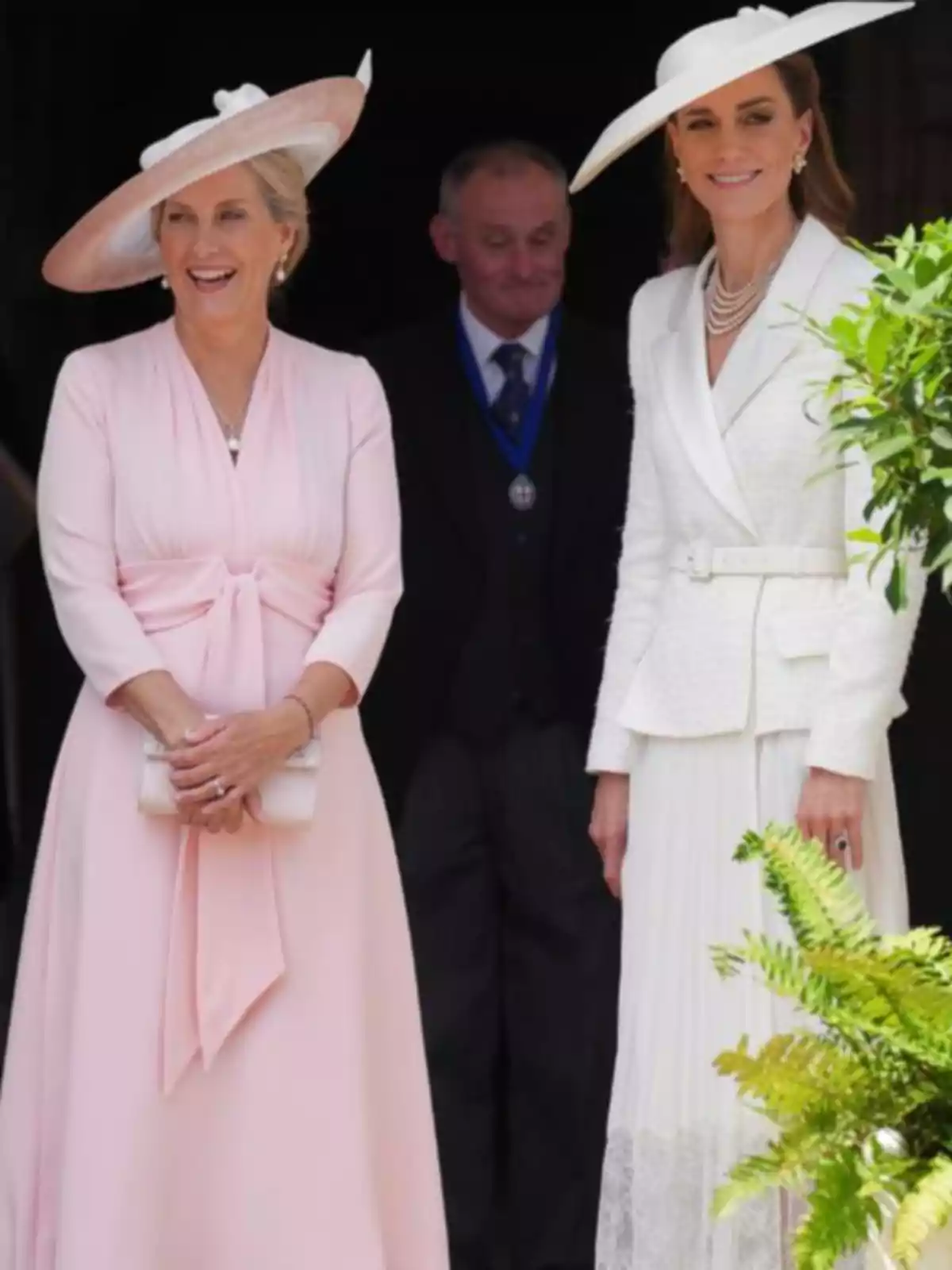 Kate Middleton y otra mujer elegantemente vestidas con sombreros claros posan sonrientes al aire libre junto a un hombre de traje en el fondo Kate Middleton y otra mujer elegantemente vestidas con sombreros claros posan sonrientes al aire libre junto a un hombre de traje en el fondo