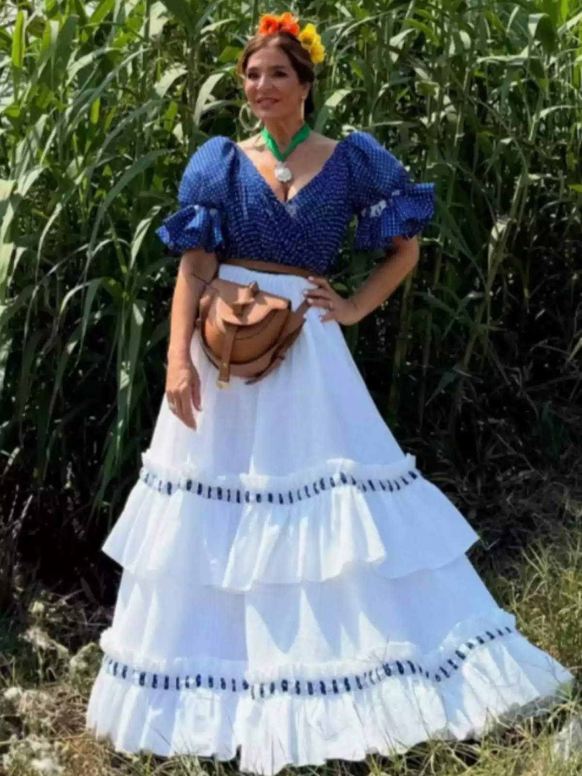 Raquel Bollo vestida con falda blanca de volantes y blusa azul con lunares, lleva flores en el cabello y una bolsa marrón en la cintura, posando al aire libre frente a plantas verdes. Raquel Bollo vestida con falda blanca de volantes y blusa azul con lunares, lleva flores en el cabello y una bolsa marrón en la cintura, posando al aire libre frente a plantas verdes.