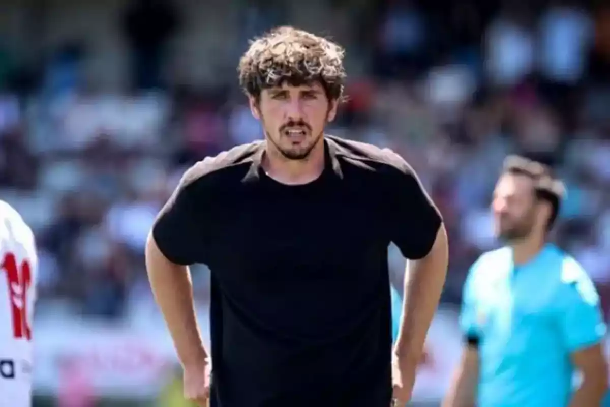 Un hombre con camiseta negra está de pie en un campo deportivo con personas desenfocadas al fondo.