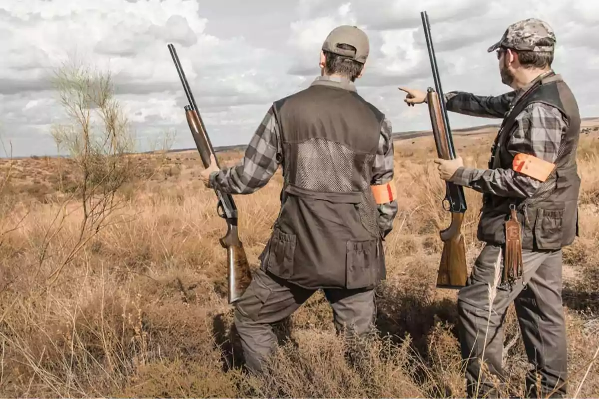 Dos cazadores con escopetas en un campo seco, uno de ellos señalando hacia la distancia.