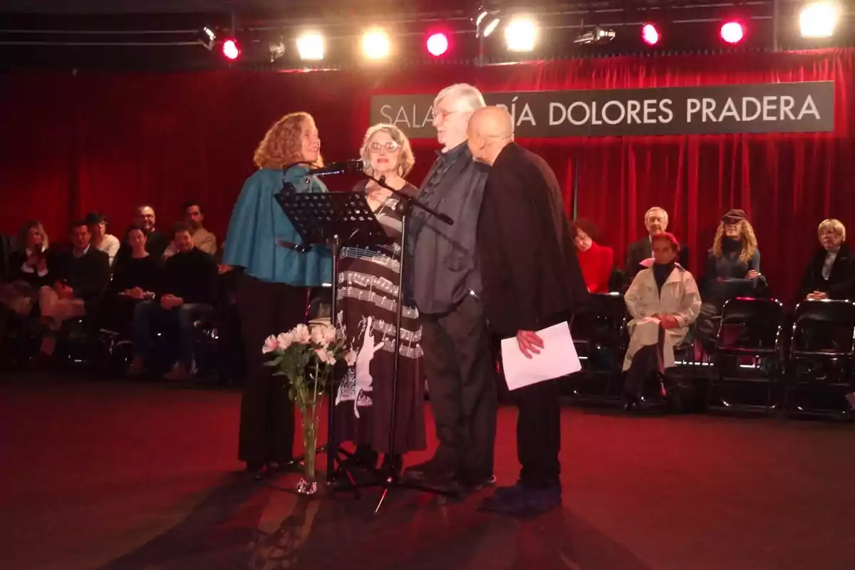 Un grupo de personas está de pie en un escenario con un micrófono, rodeados de un público sentado, en un lugar con cortinas rojas y un cartel que dice "Sala María Dolores Pradera".