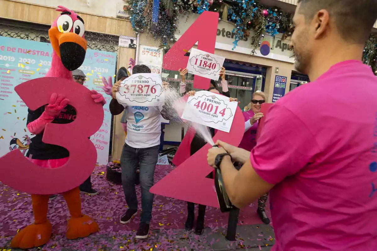 Un grupo de personas celebra con confeti y champán frente a una tienda de lotería, sosteniendo carteles con números ganadores y un disfraz de flamenco rosa.