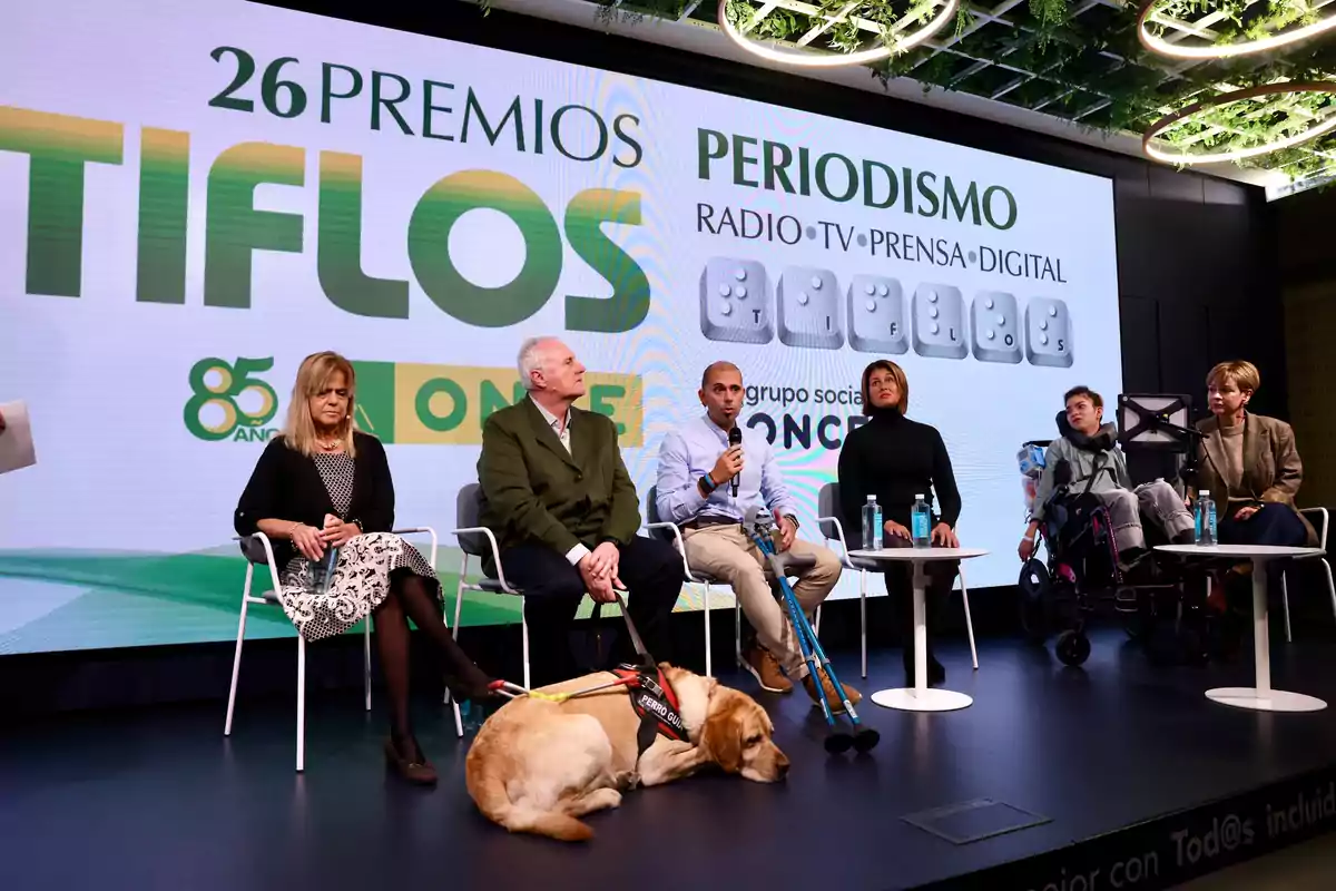 Un grupo de personas sentadas en un escenario durante la ceremonia de los 26 Premios Tiflos de Periodismo de la ONCE con un perro guía acostado en el suelo.