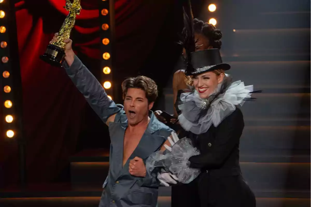 Una persona sostiene un trofeo dorado mientras otra, vestida con un atuendo elegante y un sombrero de copa, sonríe a su lado en un escenario iluminado.