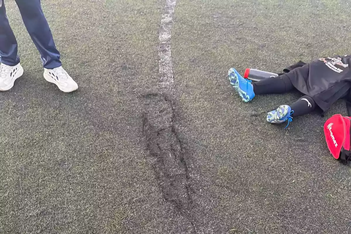 Dos personas en un campo de fútbol una de pie y otra acostada con una línea blanca en el césped.