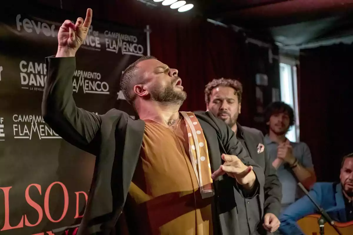 Un grupo de artistas de flamenco se presenta en un escenario, con un hombre en primer plano realizando una expresión apasionada mientras otros músicos lo acompañan en el fondo.