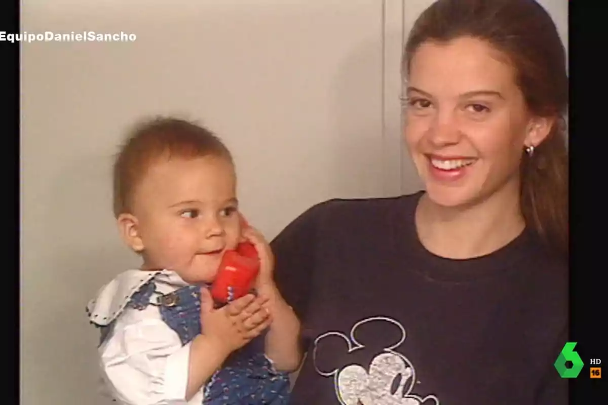 Una mujer sonriente sostiene a un bebé que juega con un teléfono de juguete rojo.