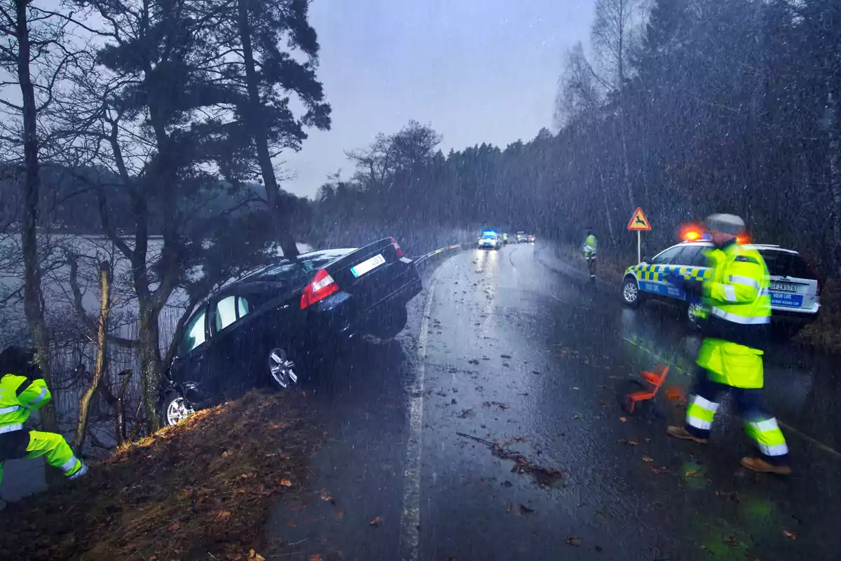 Un coche negro se ha salido de la carretera mojada y está inclinado en una zanja mientras la policía y los servicios de emergencia trabajan en el lugar bajo la lluvia.