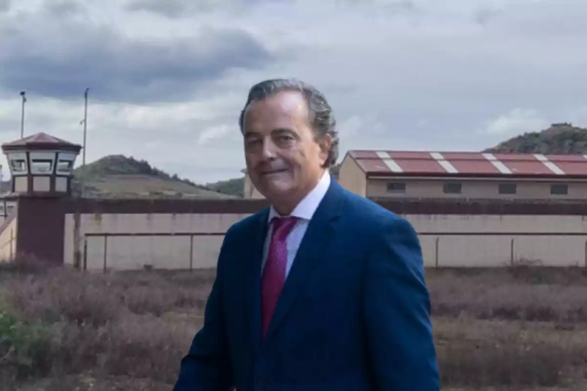 Un hombre con traje azul y corbata roja está de pie frente a un edificio con torres de vigilancia y un cielo nublado.