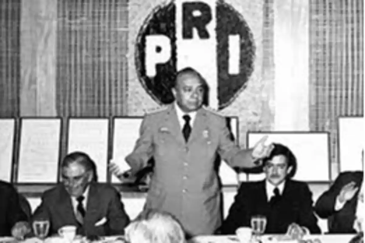 Un grupo de personas en una reunión con un hombre de pie hablando, con el logotipo del PRI en el fondo.