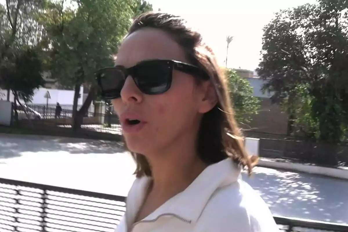 Irene Rosales con gafas de sol caminando al aire libre en un día soleado con árboles y una cerca de fondo.